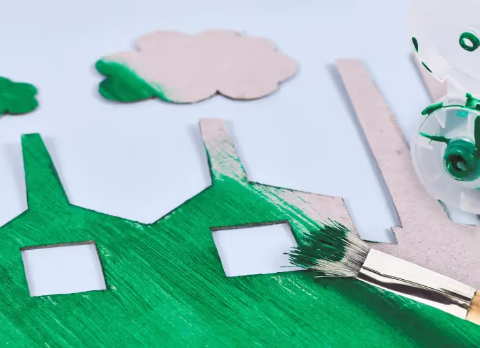 Silhouettes d'usines en carton dont une est peinte en vert au pinceau, symbole du greenwashing, sur fond bleu clair.