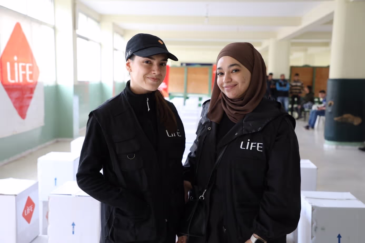 Deux jeunes femmes souriantes en veste noire LIFE posent dans une salle remplie de cartons blancs marqués du logo LIFE