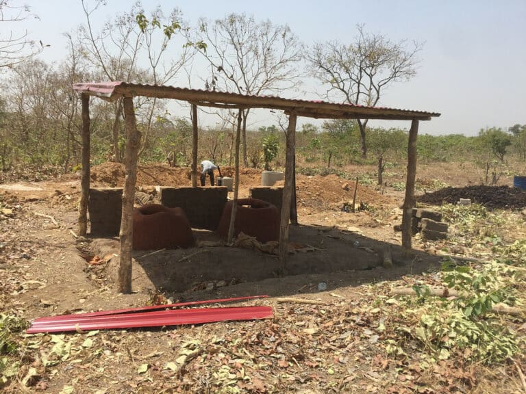 Chantier de construction de deux dômes en briques sous un abri à toit de tôle rouge, en pleine nature sèche
