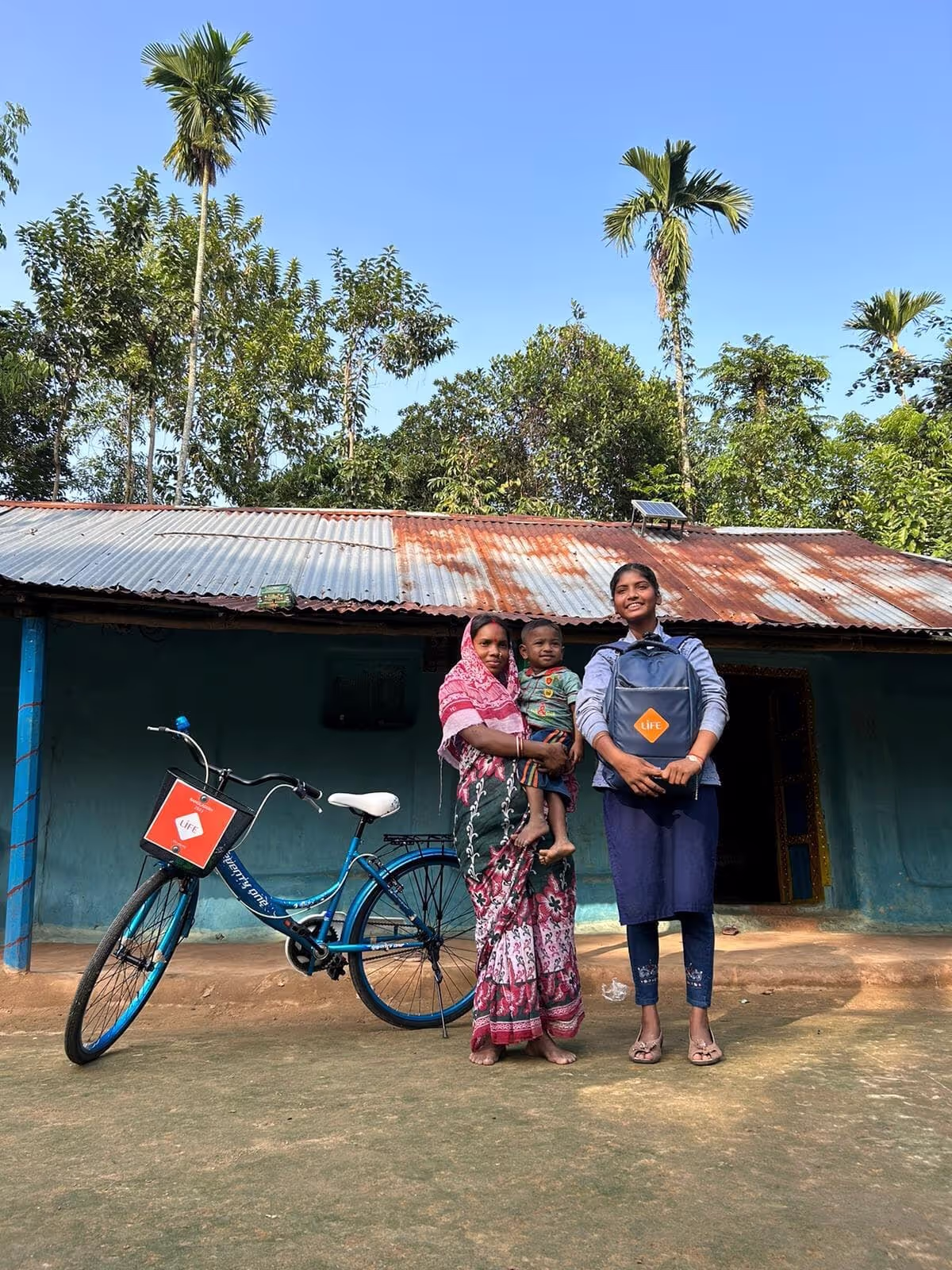 Une femme tenant un enfant et une jeune fille avec un sac à dos LIFE posent devant une maison, près d'un vélo avec panier LIFE.