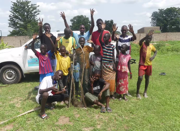 Groupe d'enfants et d'adolescents souriants qui lèvent la main, posant sur l'herbe devant un pick-up blanc, autour d'un jeune arbre planté