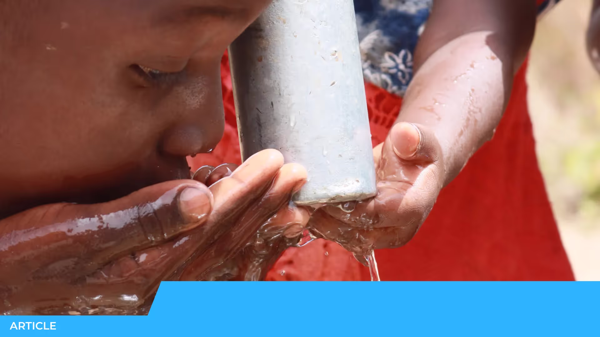 Un enfant boit au creux de ses mains l'eau qui coule d'un tuyau métallique, tenu par une autre personne en rouge