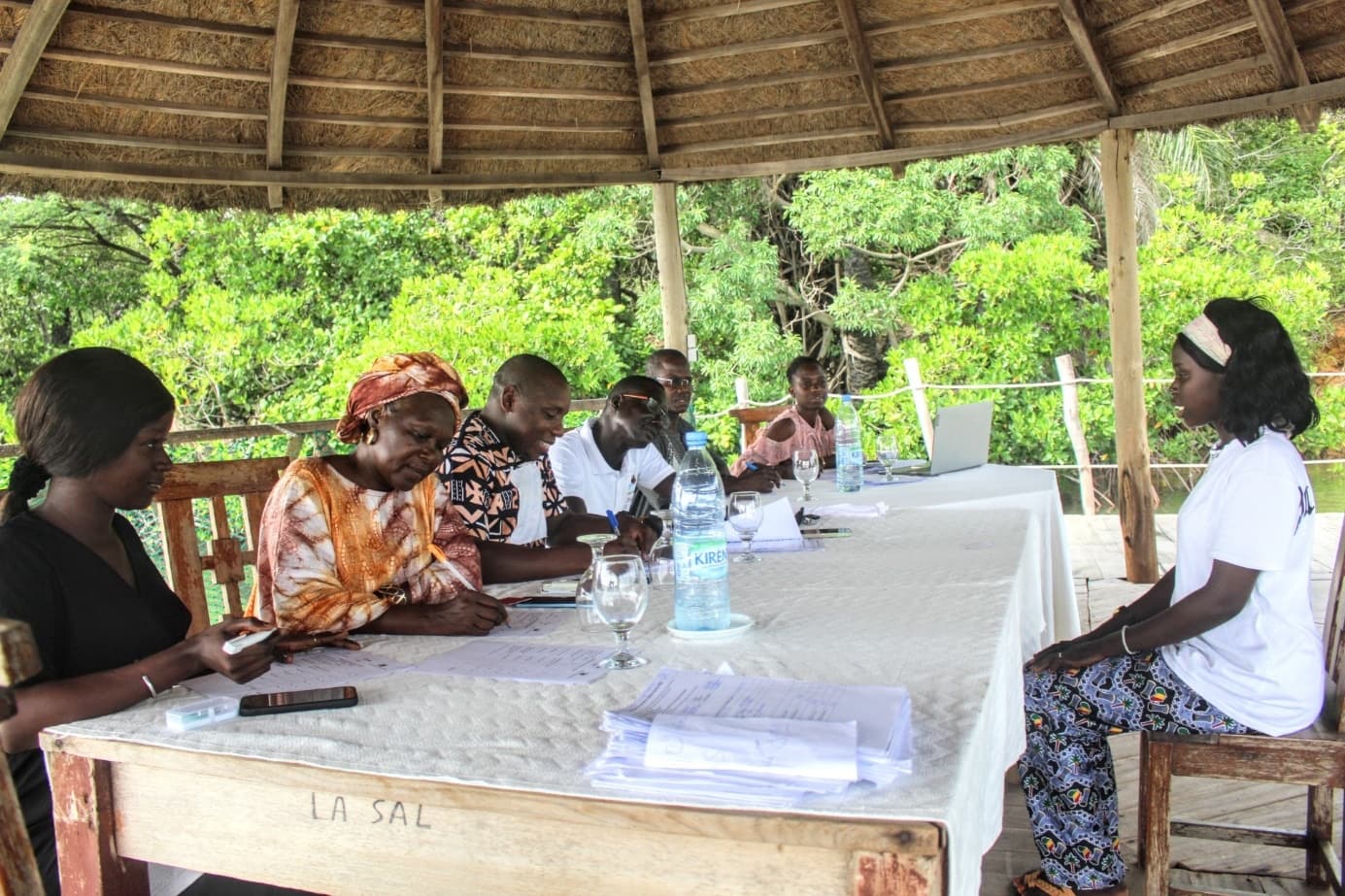 Réunion de travail autour d'une longue table sous une paillote, avec des participants qui prennent des notes face à une jeune femme